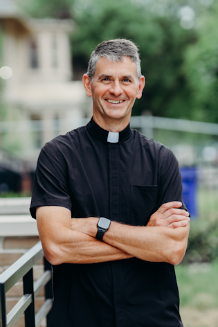 Priest wearing a black shirt with a collar, standing outdoors with arms crossed.